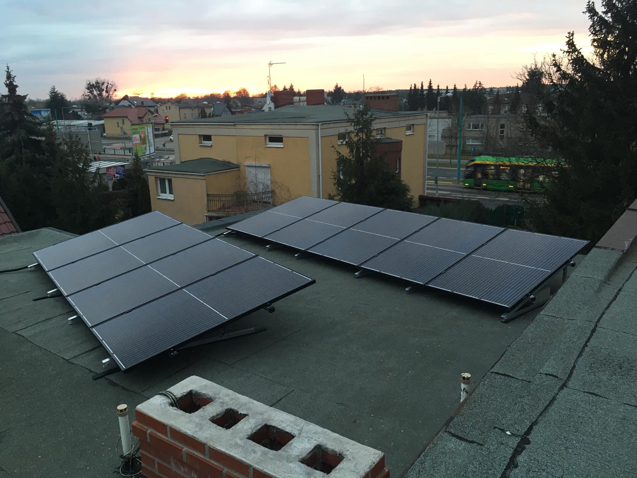 Panele słoneczne na płaskim dachu budynku mieszkalnego, widok z góry, w tle zabudowa miejska i zachodzące słońce.