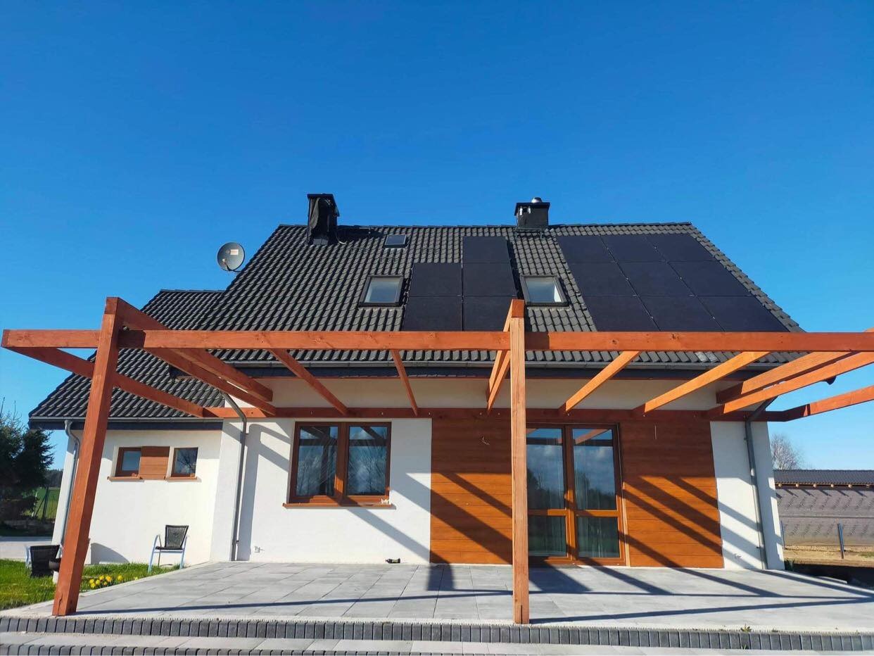 Dom jednorodzinny z panelami słonecznymi na dachu, widok z przodu, drewniana pergola nad tarasem. Uwagę zwracają ciemne panele fotowoltaiczne wkomponowane w grafitowy dachówkowy dach.