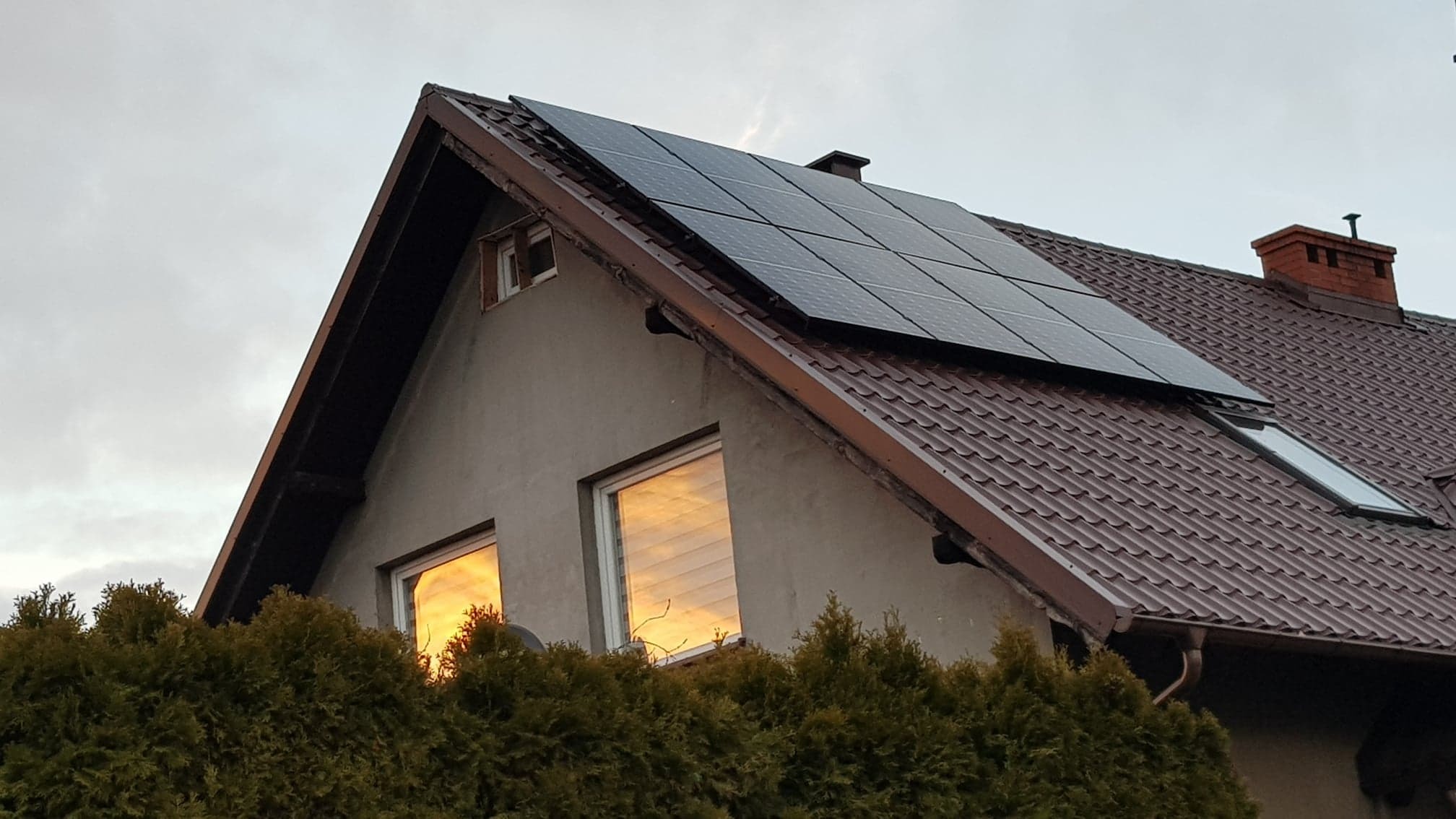 Dom jednorodzinny z brązowym dachem, na którym zamontowane są panele fotowoltaiczne; w oknach odbija się zachodzące słońce, a w tle widoczne krzewy iglaste.
