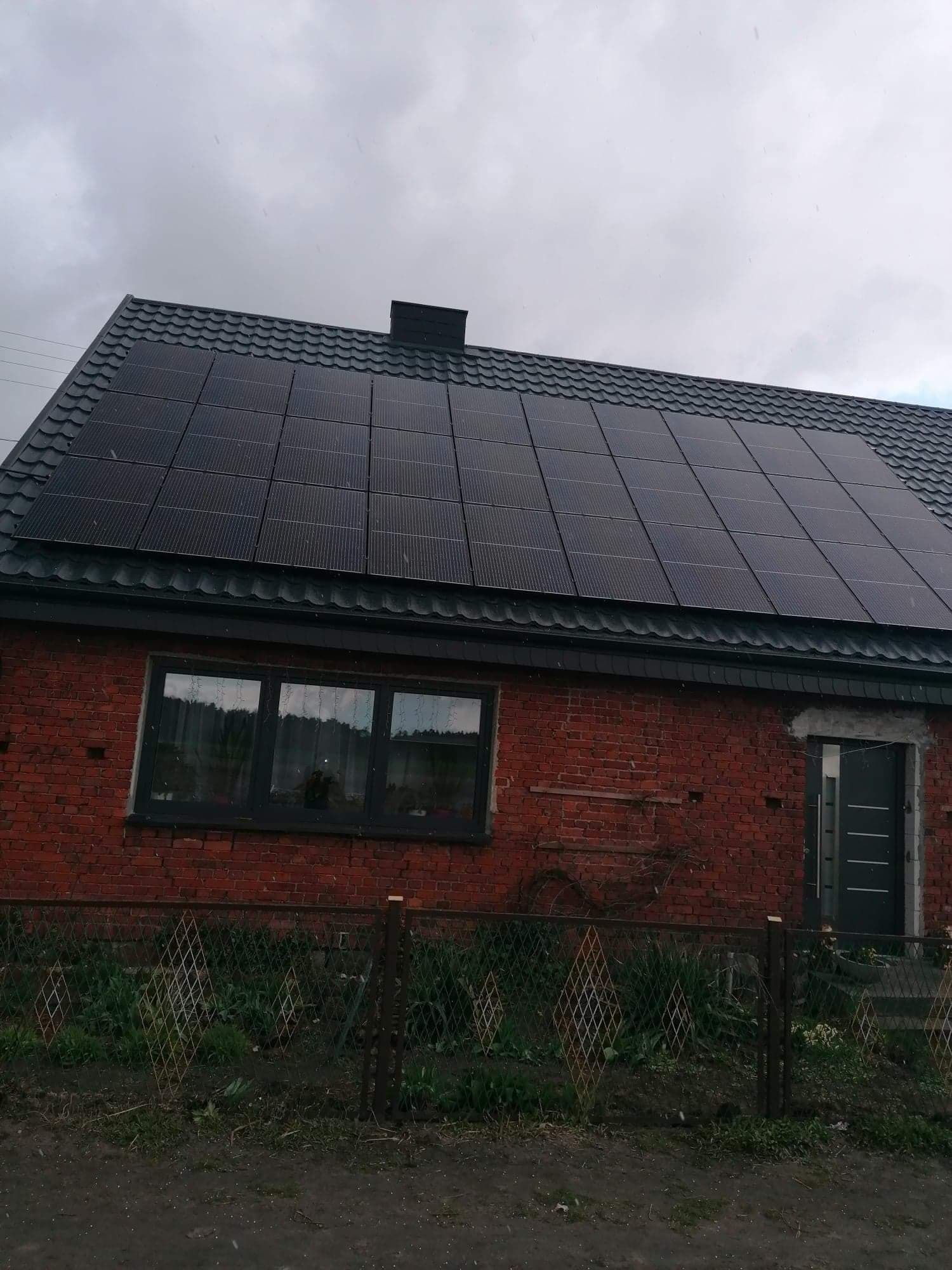 Czerwony ceglany dom z czarnymi panelami słonecznymi na dachu z metalowej dachówki i czarną stolarką okienną, widziany zza niskiego ogrodzenia.
