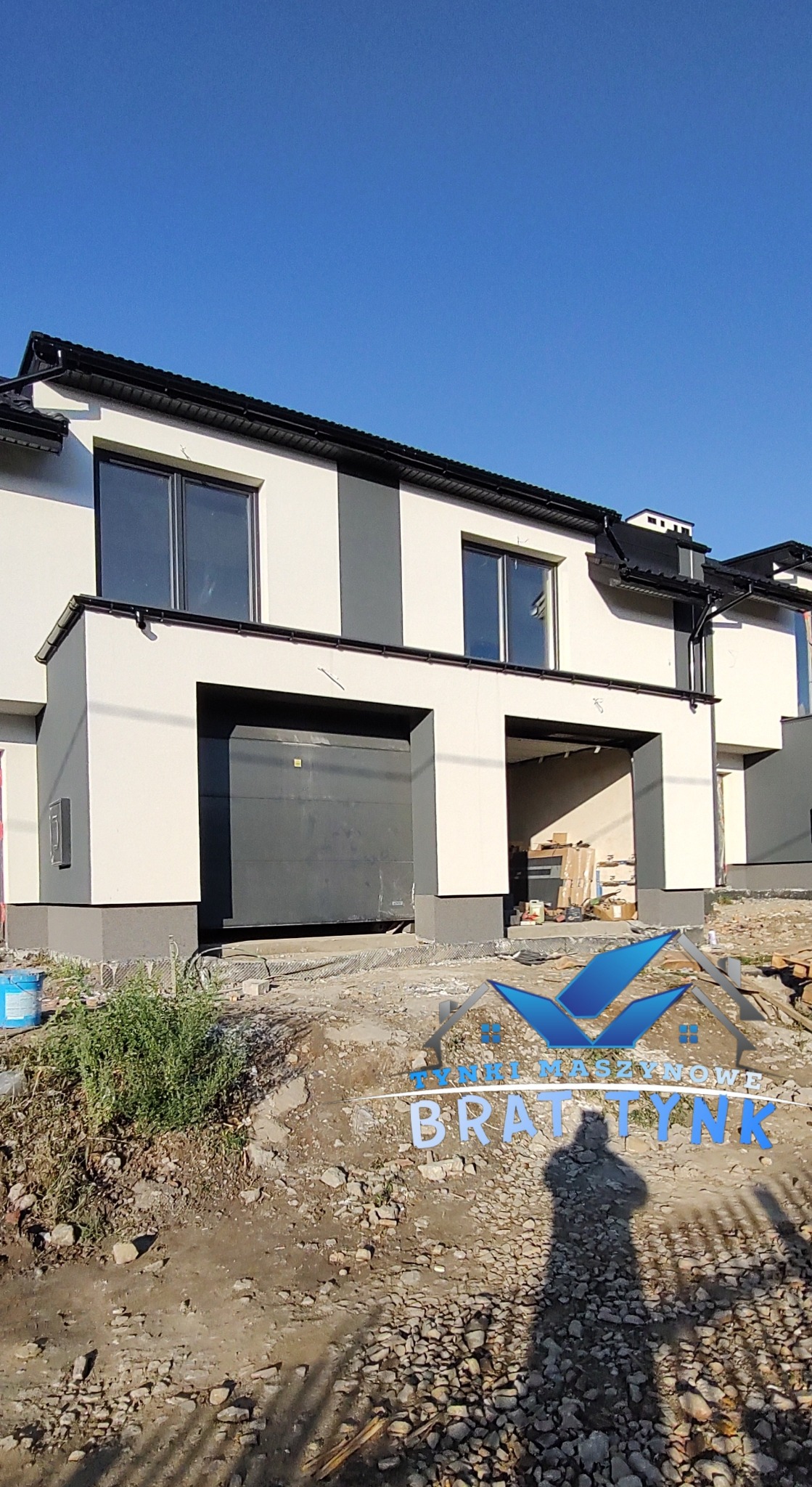 Nowo otynkowany dom w zabudowie bliźniaczej z grafitowymi akcentami, widoczny garaż i teren w trakcie prac wykończeniowych.