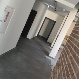 Bud-System - Korytarz w trakcie prac wykończeniowych, widoczne płytki podłogowe, białe ściany, ościeżnice drzwiowe, fragment ściany z cegły klinkierowej oraz elementy instalacji pod sufitem.