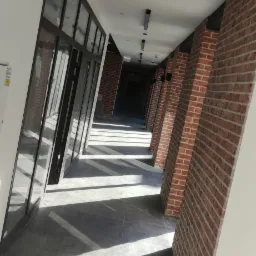 Długi korytarz z rzędami ceglanych filarów po prawej stronie i szklanymi ścianami po lewej, oświetlony promieniami słonecznymi tworzącymi geometryczne wzory na podłodze z szarych płytek.