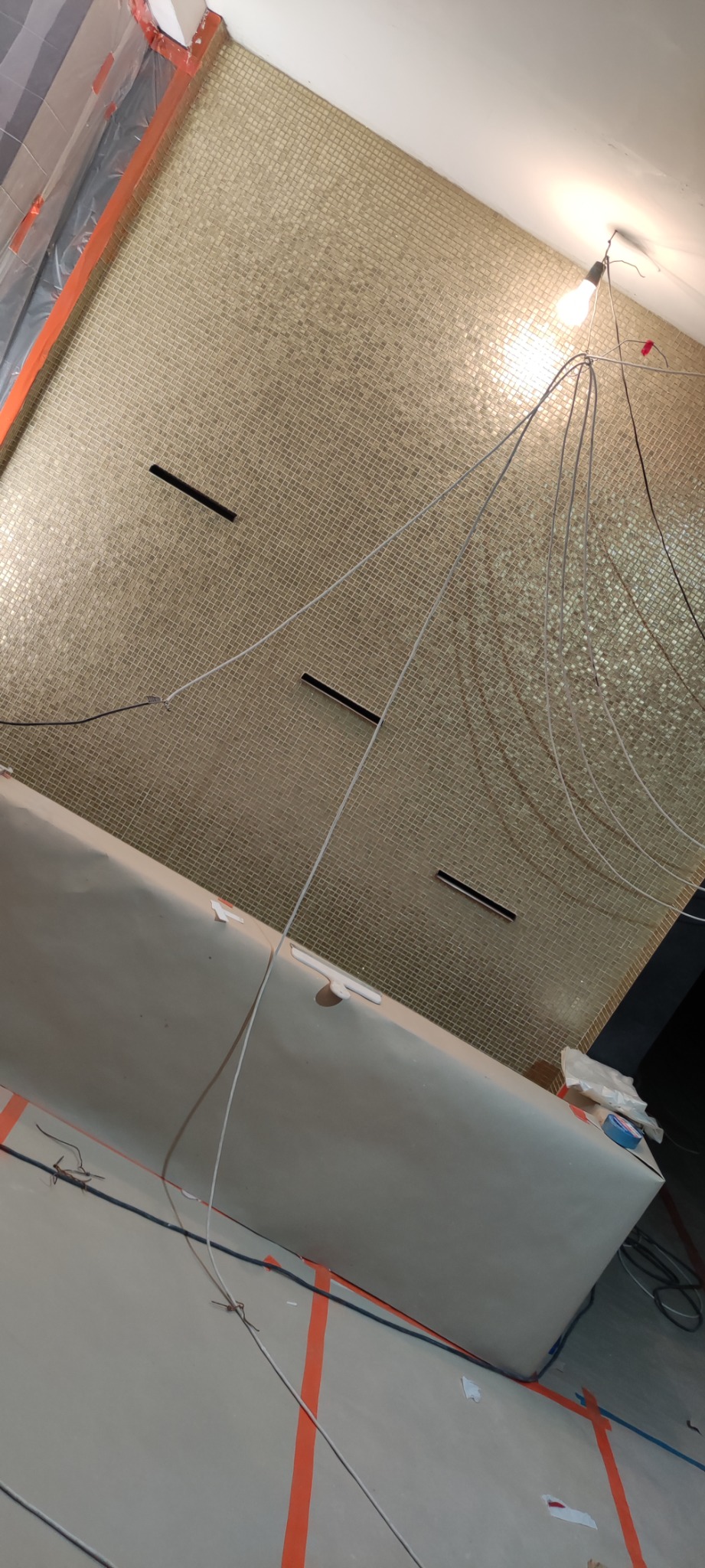 Łazienka w trakcie prac wykończeniowych: ściana wyłożona mozaiką w odcieniach beżu i złota, zabezpieczona folią podłoga, widoczne przewody elektryczne i oświetlenie tymczasowe.