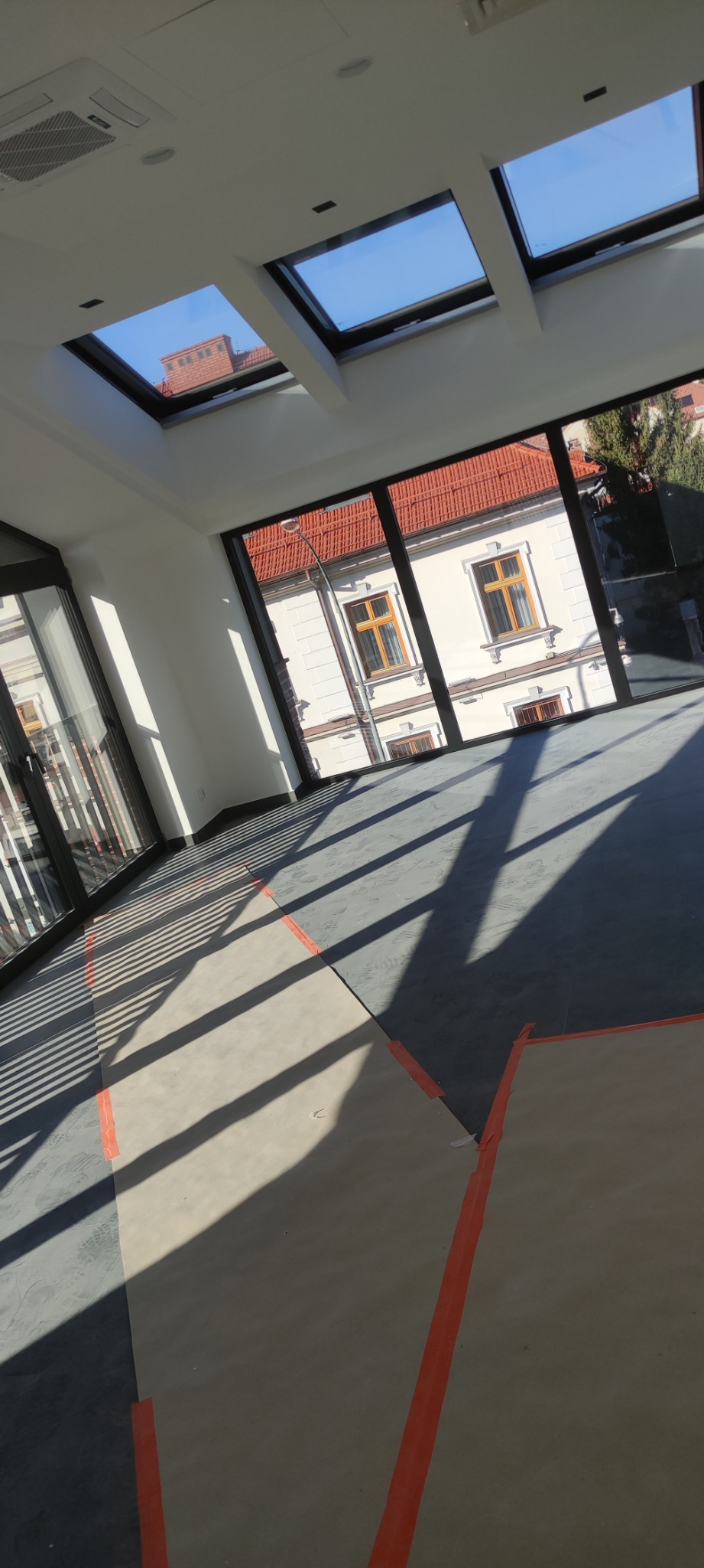 Wnętrze w trakcie prac przygotowawczych do wykończenia podłogi, zabezpieczone kartonem i taśmą malarską, widoczne okna dachowe i duże okna z widokiem na budynek z czerwonym dachem.