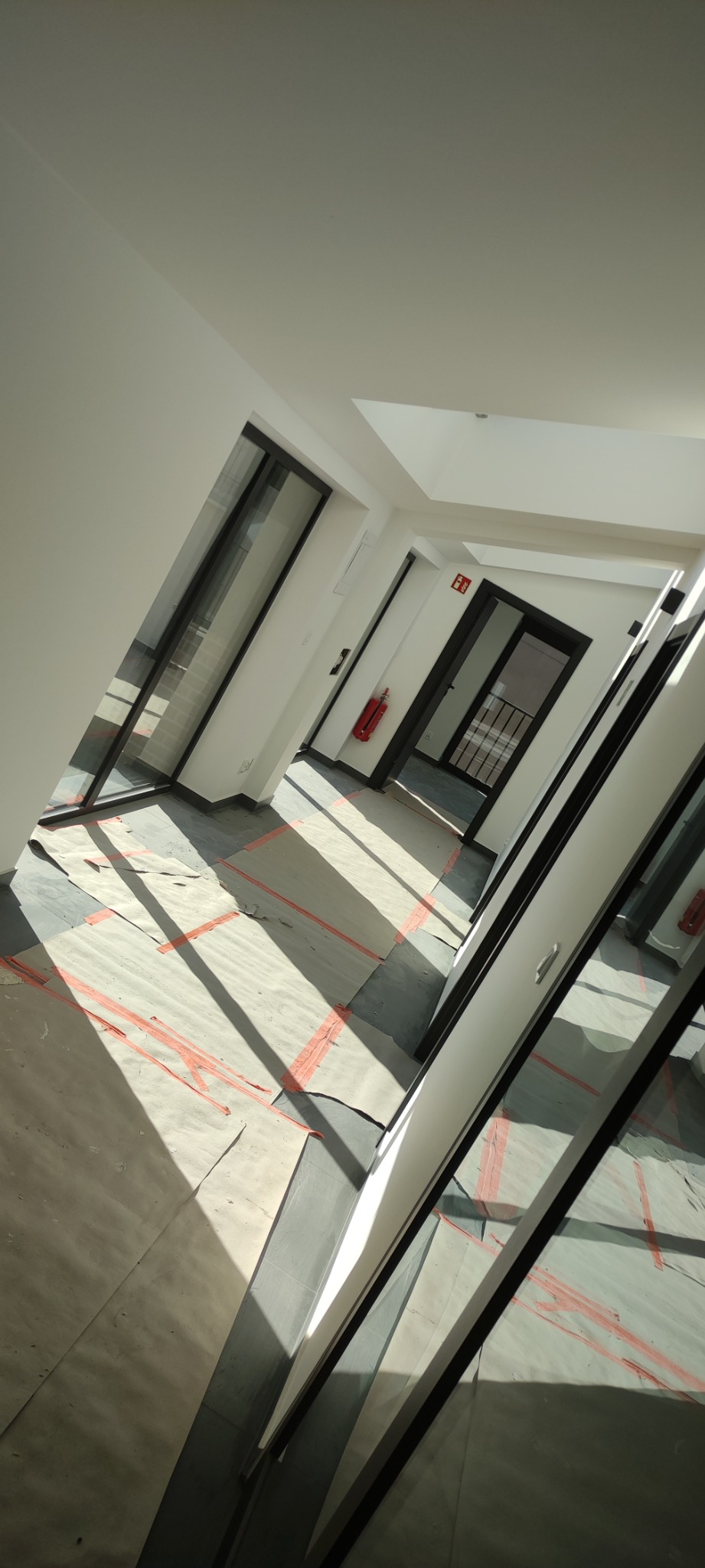 Korytarz w trakcie prac wykończeniowych, podłoga zabezpieczona kartonem i taśmą, widoczne duże okna z czarnymi ramami i białe ściany, w głębi drzwi przeciwpożarowe i gaśnica.
