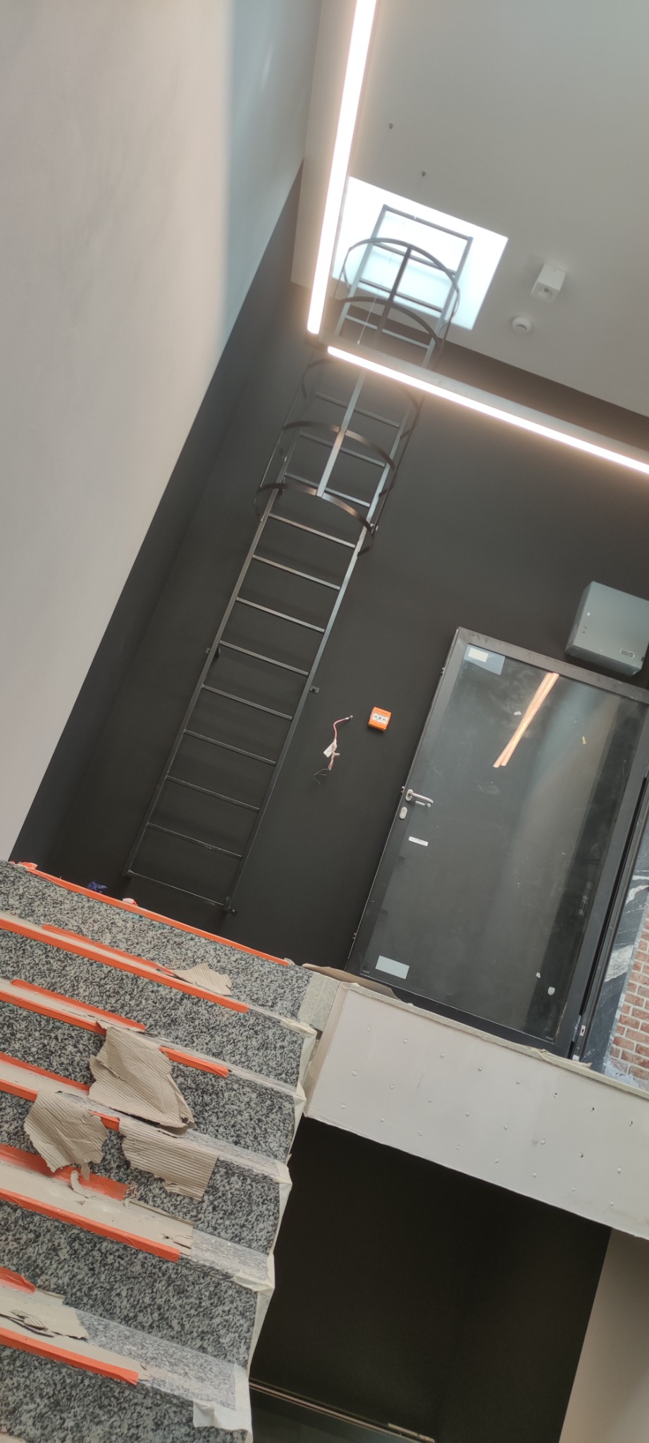 Wnętrze klatki schodowej w trakcie wykańczania: zabezpieczone schody z szarego lastryko z pomarańczowymi paskami, czarna ściana z metalową drabiną ewakuacyjną prowadzącą do świetlika, drzwi...