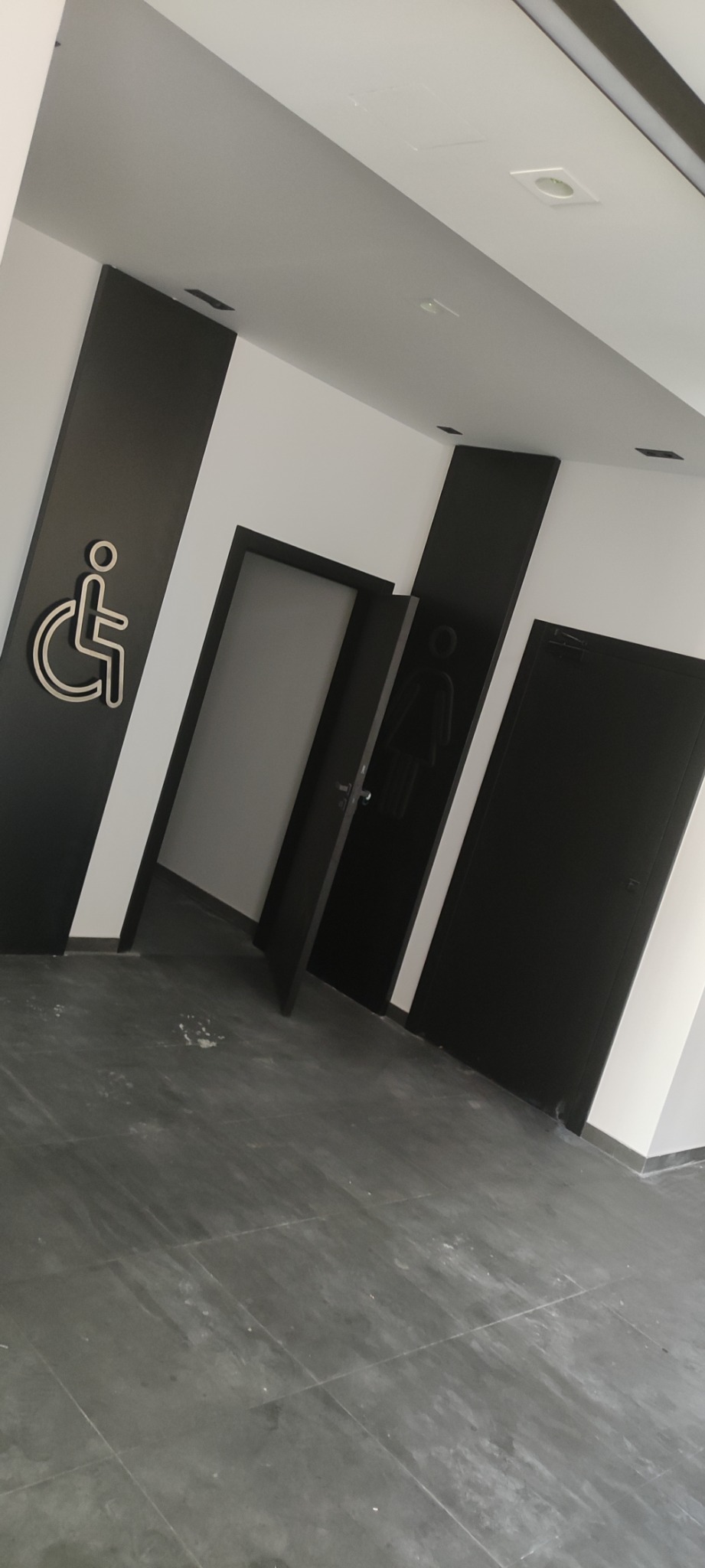 Wnętrze publicznej toalety z czarnymi drzwiami i oznaczeniami dla osób niepełnosprawnych i kobiet na czarnych panelach, widoczne szare płytki podłogowe.