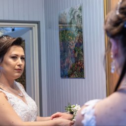 Fotograf Polkowski - Panna młoda w białej sukni z koronkowymi zdobieniami, spoglądająca w lustro, z widocznym bukiecikiem na nadgarstku i ozdobą we włosach.