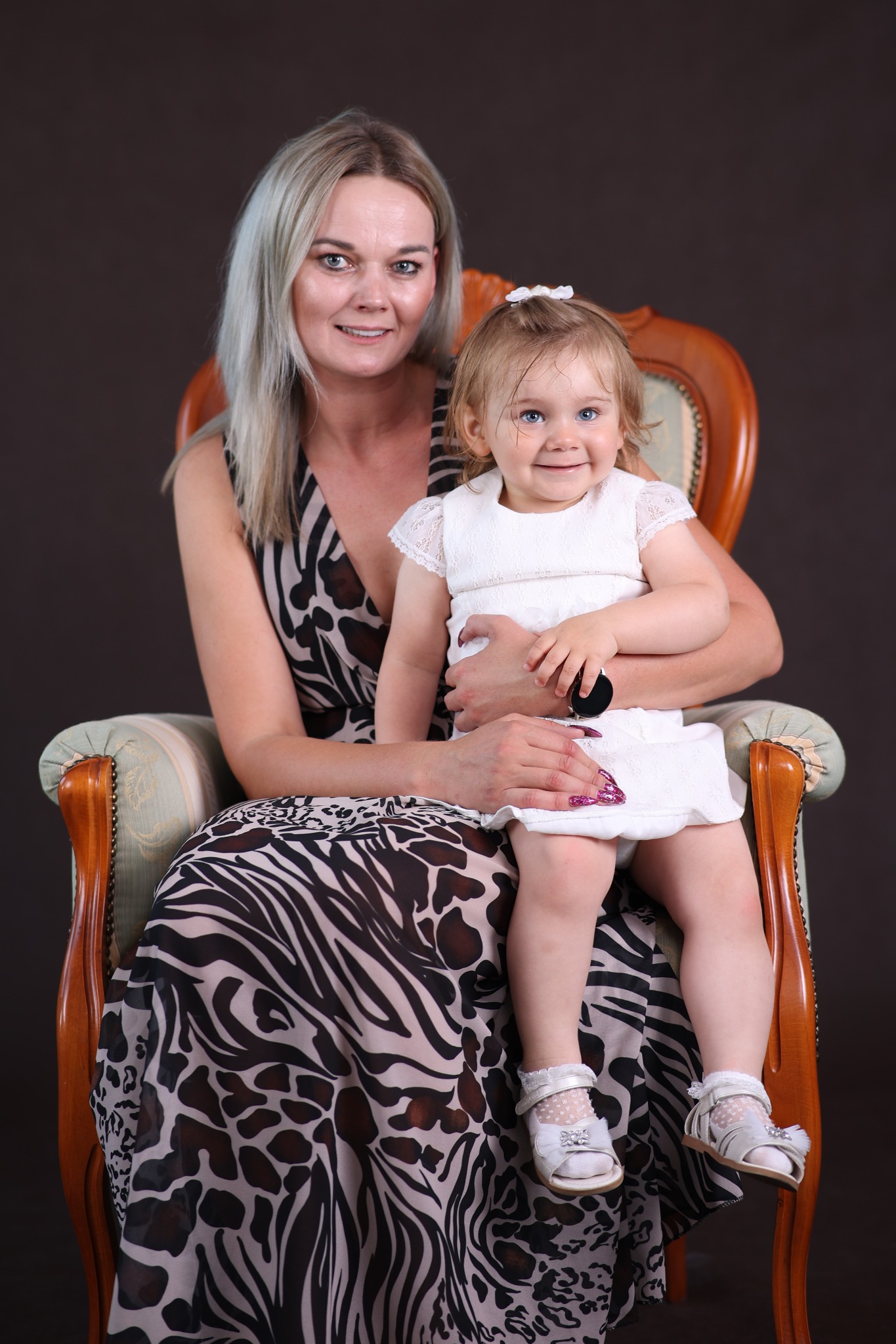 Portret matki z córeczką na kolanach w fotelu, ciemne tło, dziewczynka w białej sukience i sandałkach, kobieta w sukience w zwierzęcy wzór, uśmiechnięte twarze.