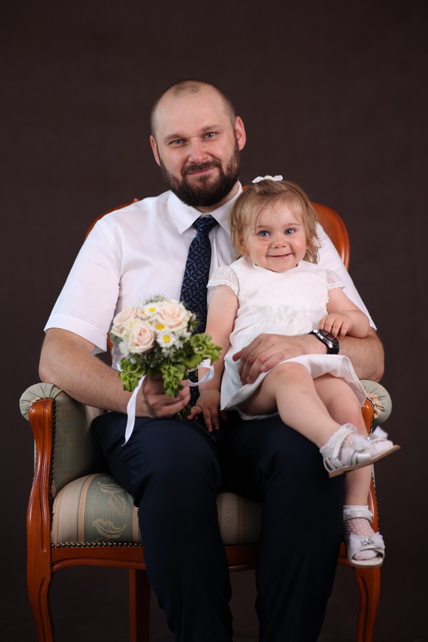 Uśmiechnięty mężczyzna w koszuli i krawacie siedzi w fotelu, trzymając na kolanach ubraną na biało dziewczynkę z bukietem róż i margaretek.