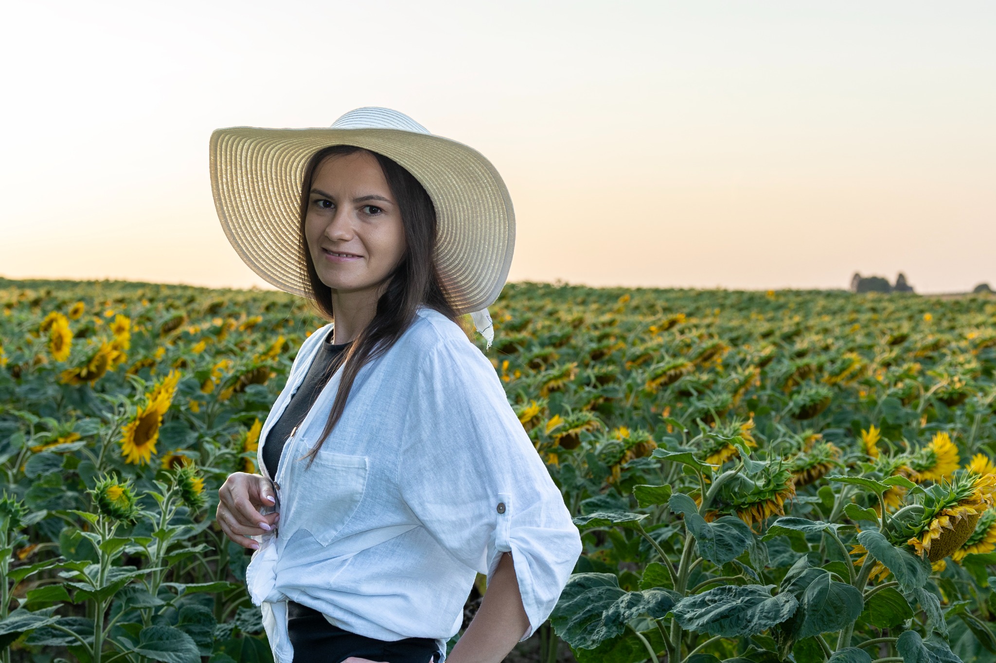 Kobieta w kapeluszu i białej koszuli na tle pola słoneczników o zachodzie słońca, z delikatnym uśmiechem i ręką opartą na biodrze.