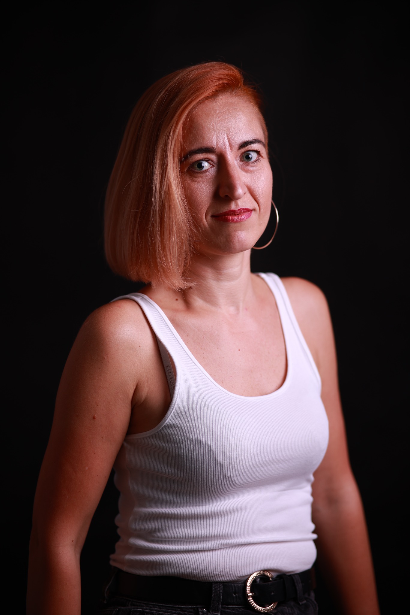 Portret kobiety z rudymi włosami, w białym topie na tle czarnego tła, z kolczykiem kołem i paskiem z ozdobną klamrą.