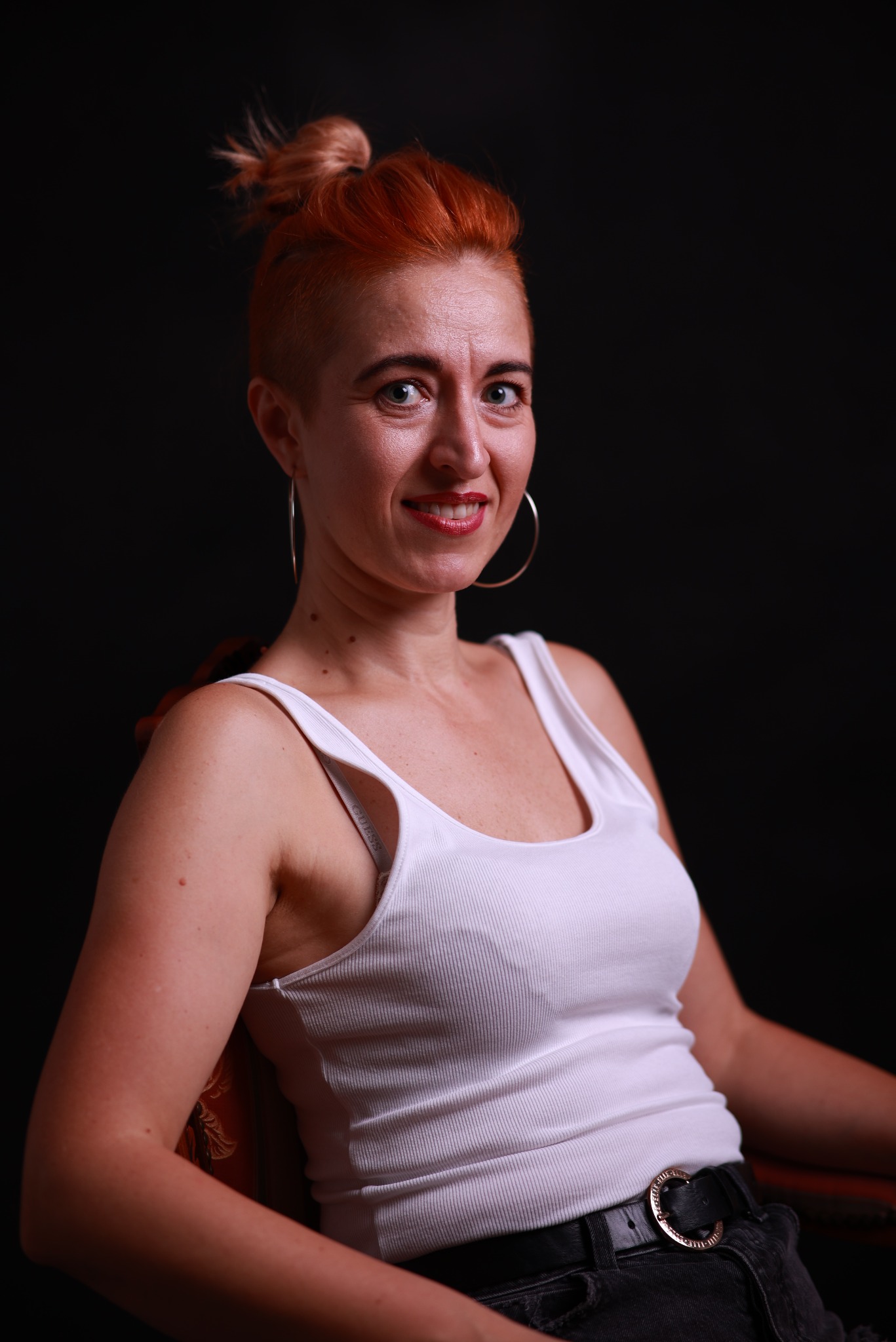 Portret uśmiechniętej kobiety z rudymi włosami upiętymi w kok, w białym podkoszulku na tle czarnego tła.