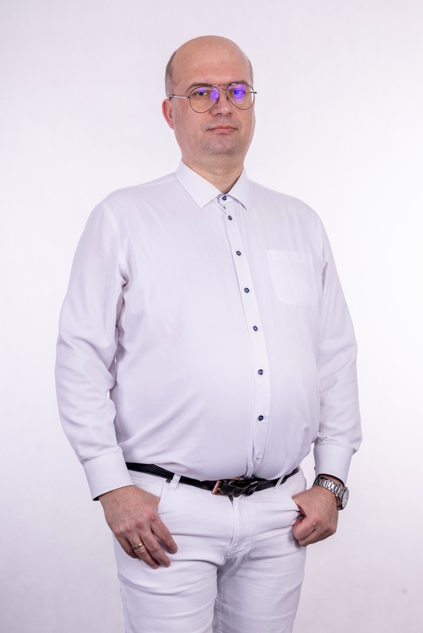 Portret mężczyzny w białej koszuli i spodniach na jasnym tle, wykonany w Bełchatowie, z widocznym zegarkiem na ręce i okularami z odblaskami.