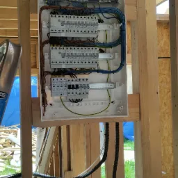 Rozdzielnica elektryczna zamontowana w drewnianej konstrukcji szkieletowej budynku w trakcie budowy, widoczne liczne kable w peszlach i przewody podłączone do bezpieczników i listew zaciskowych.