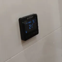 Czarny regulator temperatury Acurel zamontowany na jasnej, błyszczącej ścianie, wyświetlający godzinę 00:00, temperaturę zadaną 15 stopni Celsjusza i temperaturę pokojową 21 stopni Celsjusza.