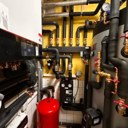 Firma Handlowo-Usługowa Robert Polis (Gotowa Chata) - Kotłownia z kotłem Viessmann, zbiornikiem akumulacyjnym i rozbudowaną instalacją hydrauliczną z licznymi zaworami, manometrem i zabezpieczeniami, w pomieszczeniu z żółtymi ścianami i białymi...