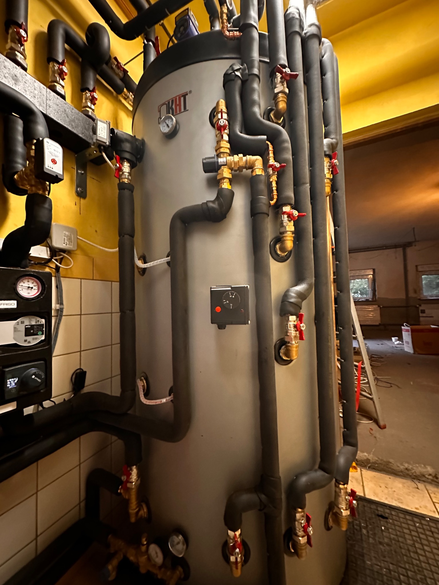 Szary zbiornik akumulacyjny KHT z licznymi rurami, zaworami i izolacją termiczną w pomieszczeniu z żółtymi ścianami i białą glazurą. Widoczny fragment instalacji grzewczej i kontrolnej.