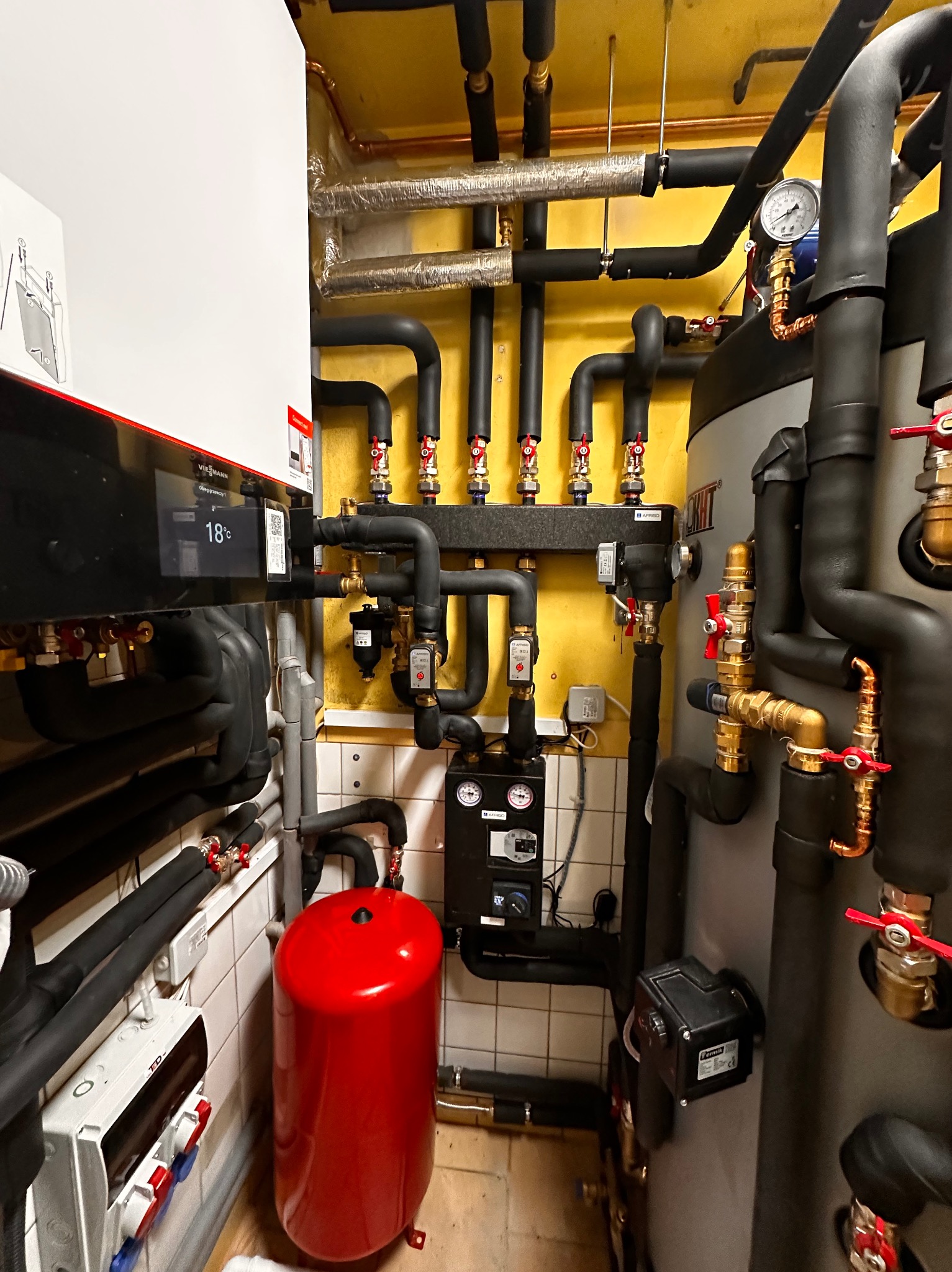 Kotłownia z kotłem Viessmann, zbiornikiem akumulacyjnym i rozbudowaną instalacją hydrauliczną z licznymi zaworami, manometrem i zabezpieczeniami, w pomieszczeniu z żółtymi ścianami i białymi...