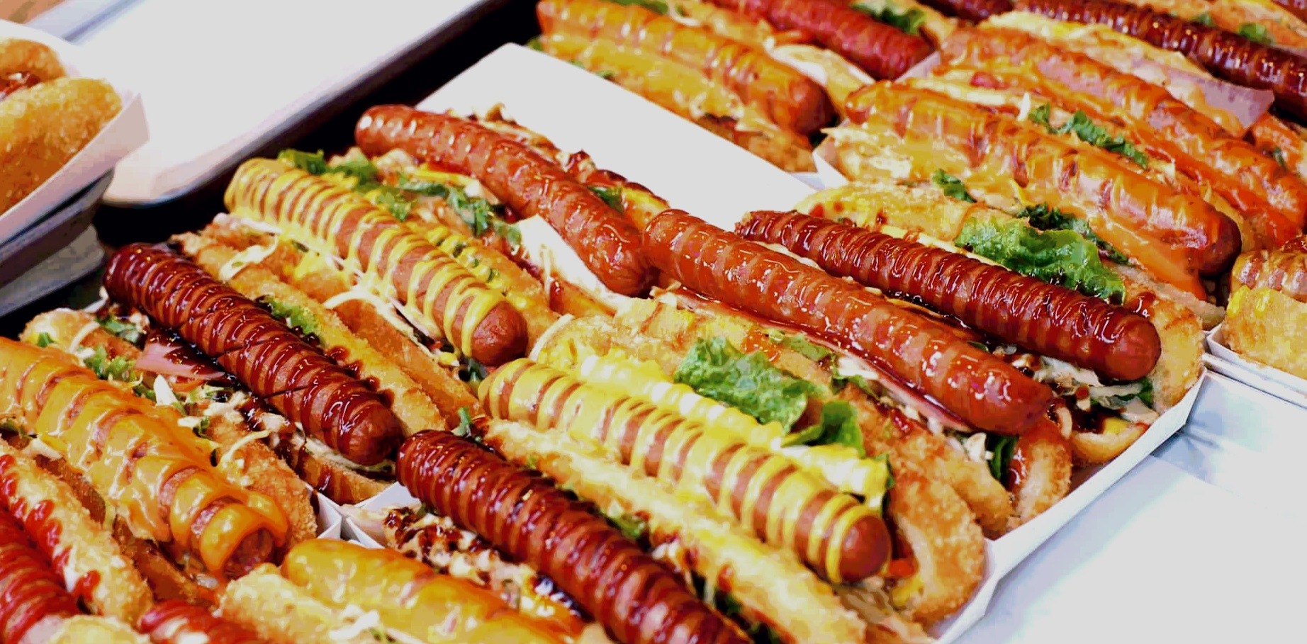 Rządki apetycznie wyglądających hot dogów z różnymi sosami, w tym musztardą, ketchupem i sosem BBQ, ułożonych na białych tackach, gotowych do podania.