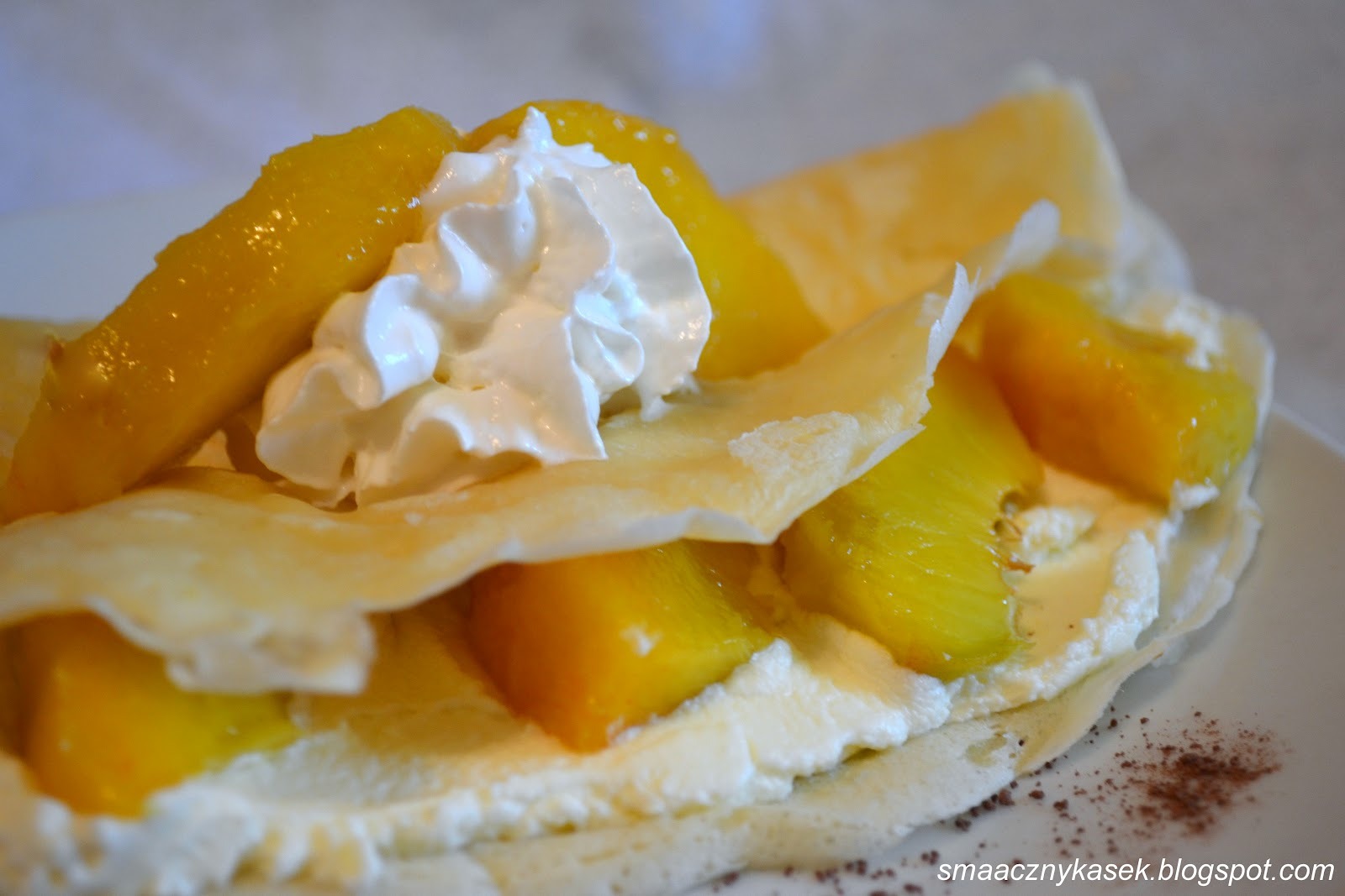 Delikatny naleśnik z kremem, kawałkami mango i bitą śmietaną, posypany delikatnie kakao na białym talerzu.