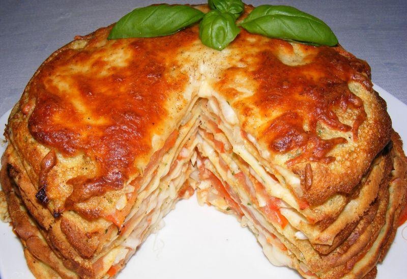 Warstwowy tort naleśnikowy z pomidorami i serem, udekorowany liśćmi bazylii, z wyciętym kawałkiem ukazującym wnętrze.