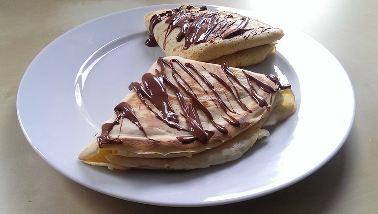 Dwa trójkątne naleśniki na białym talerzu, polane ciemną, płynną czekoladą, z widocznym jasnym nadzieniem.