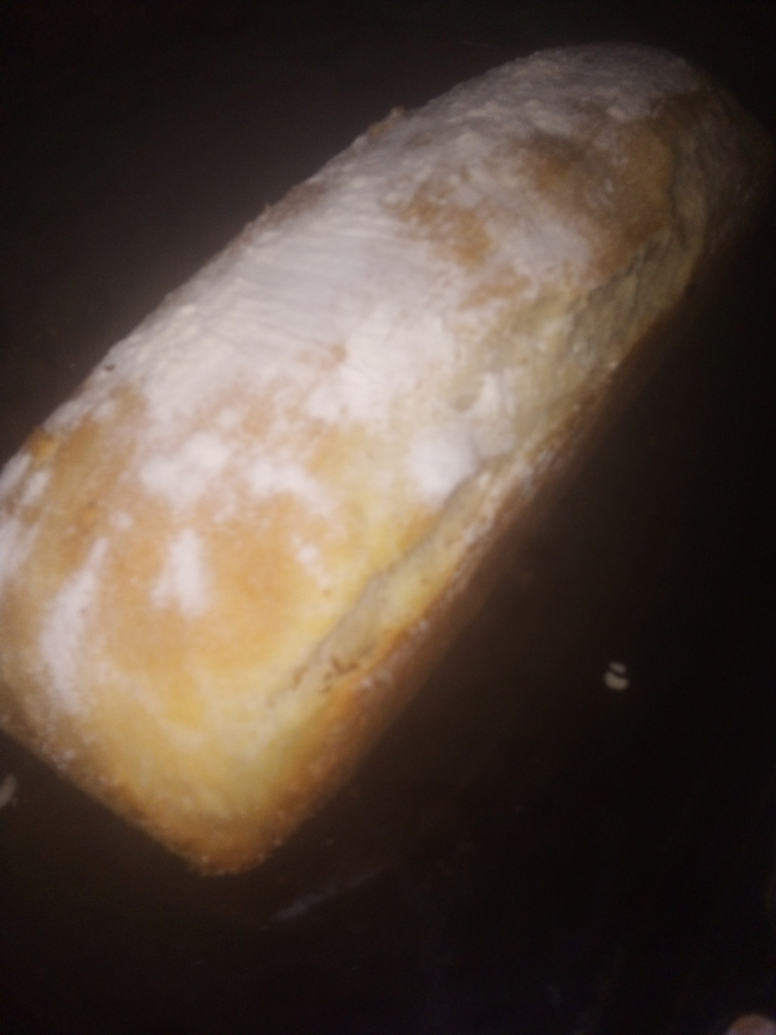 Podłużny bochenek chleba posypany mąką, leżący na ciemnym tle.