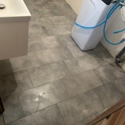 W&B bau-wiel - Ułożona szara, matowa podłoga z płytek w łazience, widoczny fragment umywalki i zmiękczacz wody podłączony niebieskimi wężami.