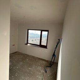 W&B bau-wiel - Pokój w trakcie szpachlowania ścian, widoczna szlifierka do gładzi na wysięgniku, okno z widokiem na zewnątrz, podłoga zabezpieczona przed zabrudzeniem.