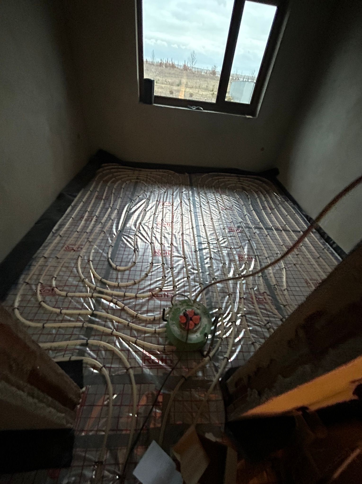 Montaż ogrzewania podłogowego w pomieszczeniu z oknem dachowym, widoczne rury grzewcze ułożone na siatce izolacyjnej, zielony rozdzielacz i fragment schodów.