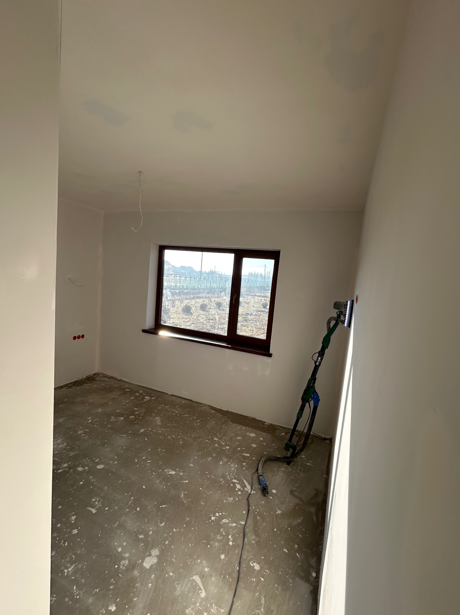 Pokój w trakcie szpachlowania ścian, widoczna szlifierka do gładzi na wysięgniku, okno z widokiem na zewnątrz, podłoga zabezpieczona przed zabrudzeniem.
