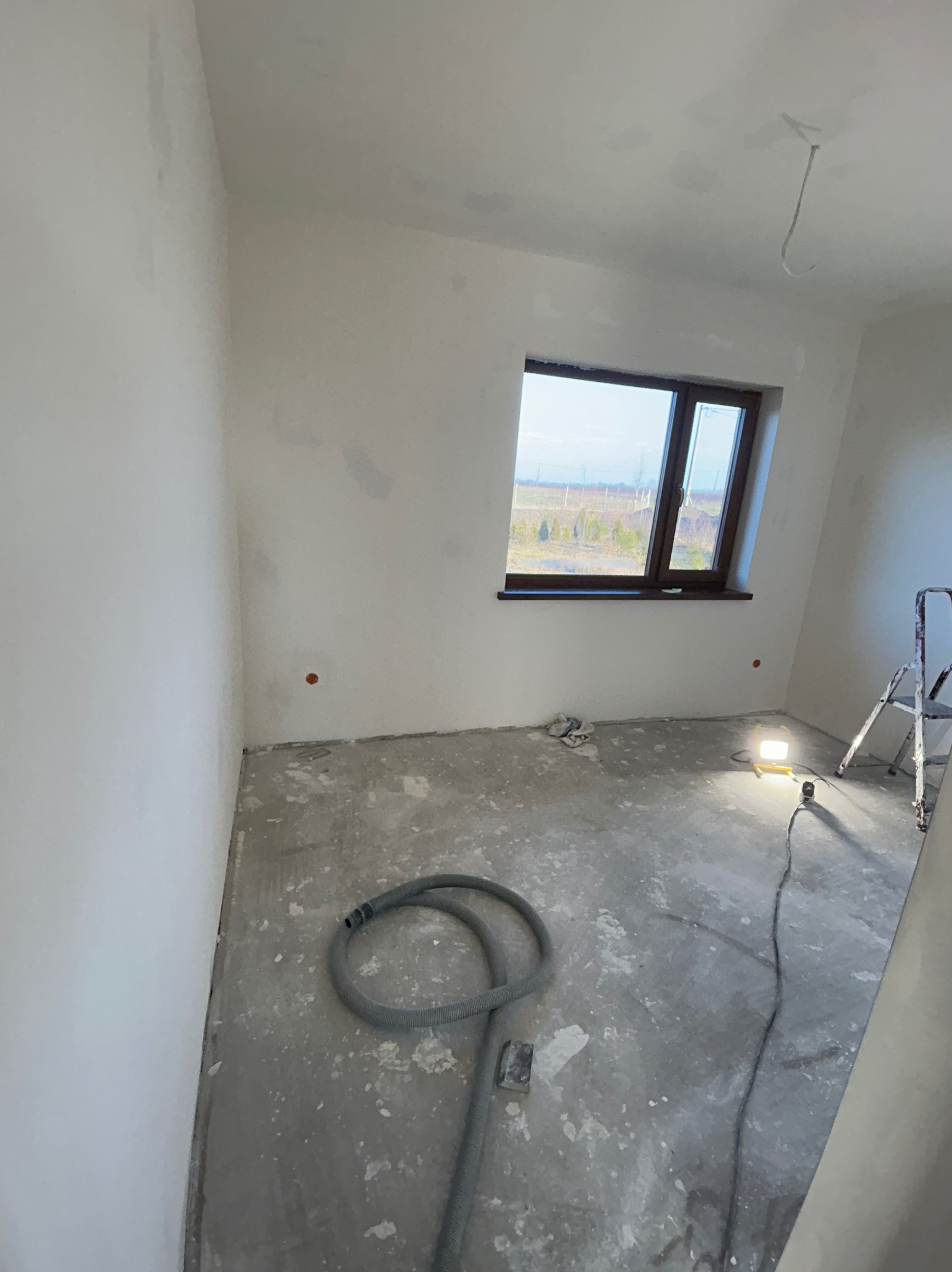 Pokój w trakcie prac wykończeniowych: ściany po gruntowaniu, widoczne ślady po szpachlowaniu, na podłodze wąż od odkurzacza przemysłowego, drabina i lampa budowlana.