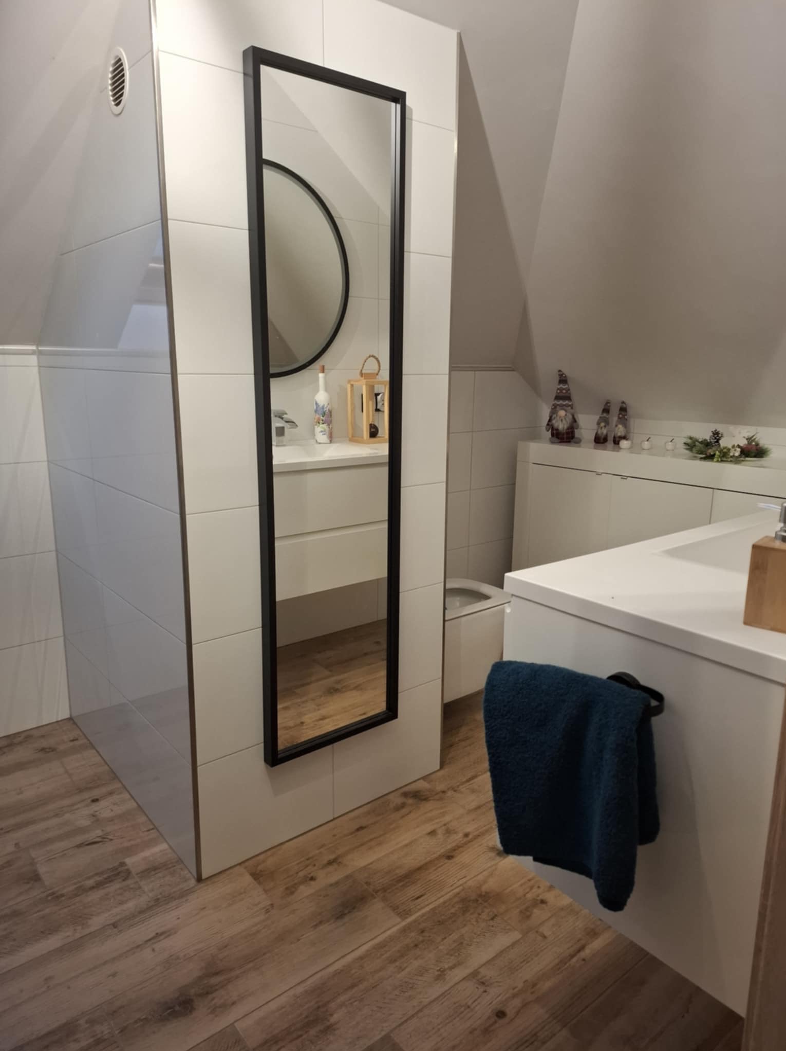 Ukończona łazienka na poddaszu z drewnopodobną podłogą, białą armaturą i grzejnikiem drabinkowym oraz wysokim lustrem w czarnej ramie.