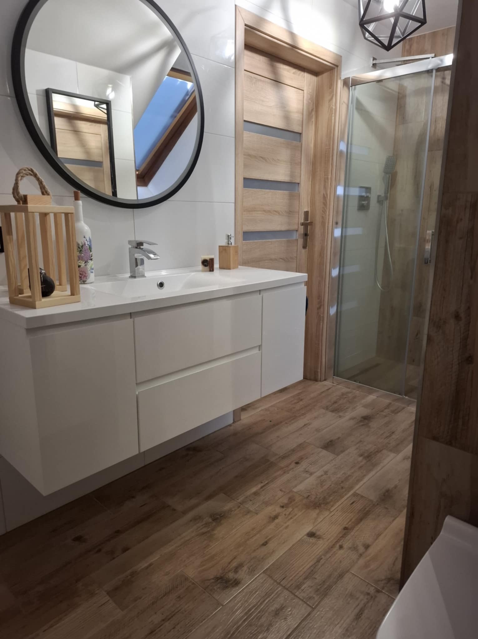 Nowoczesna łazienka z drewnopodobną podłogą, białymi płytkami i okrągłym lustrem, kabina prysznicowa ze szklanymi drzwiami i drewniana szafka pod umywalką.