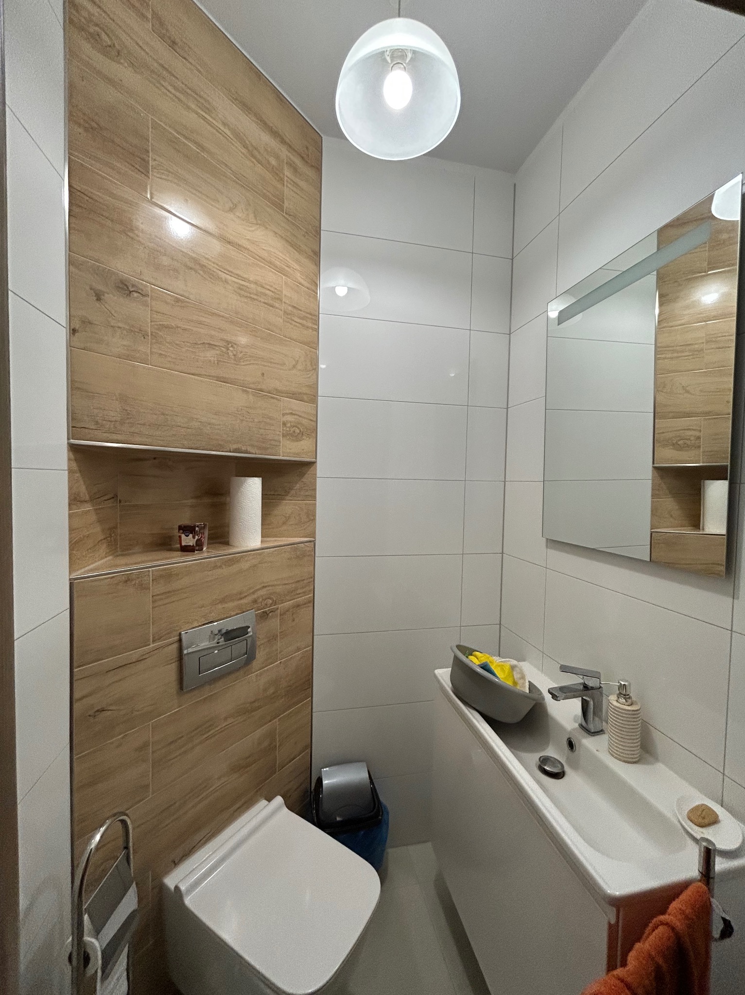 Nowoczesna łazienka z drewnopodobnymi płytkami na ścianie, białą armaturą, umywalką wpuszczaną w blat i lustrem z wbudowaną szafką, oświetlona wiszącą lampą.