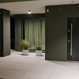 CUBE OGRODY JAKUB SZYMAŃSKI - Nowoczesne wejście do domu z minimalistycznym ogrodem: dwie białe donice z trawami ozdobnymi na białym żwirze, szara elewacja i drzwi wejściowe, oświetlenie punktowe.