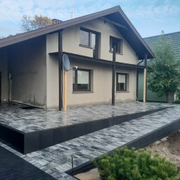 CUBE OGRODY JAKUB SZYMAŃSKI - Nowoczesny taras z szarej kostki brukowej ułożonej dwupoziomowo przed domem z brązowym dachem, widoczna antena satelitarna na elewacji budynku.