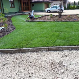 Mężczyzna w kucki podlewa świeżo założony trawnik z automatycznym systemem nawadniania, widoczne obrzeża z kostki, rabata z korą i nasadzeniami oraz żwirowy podjazd.