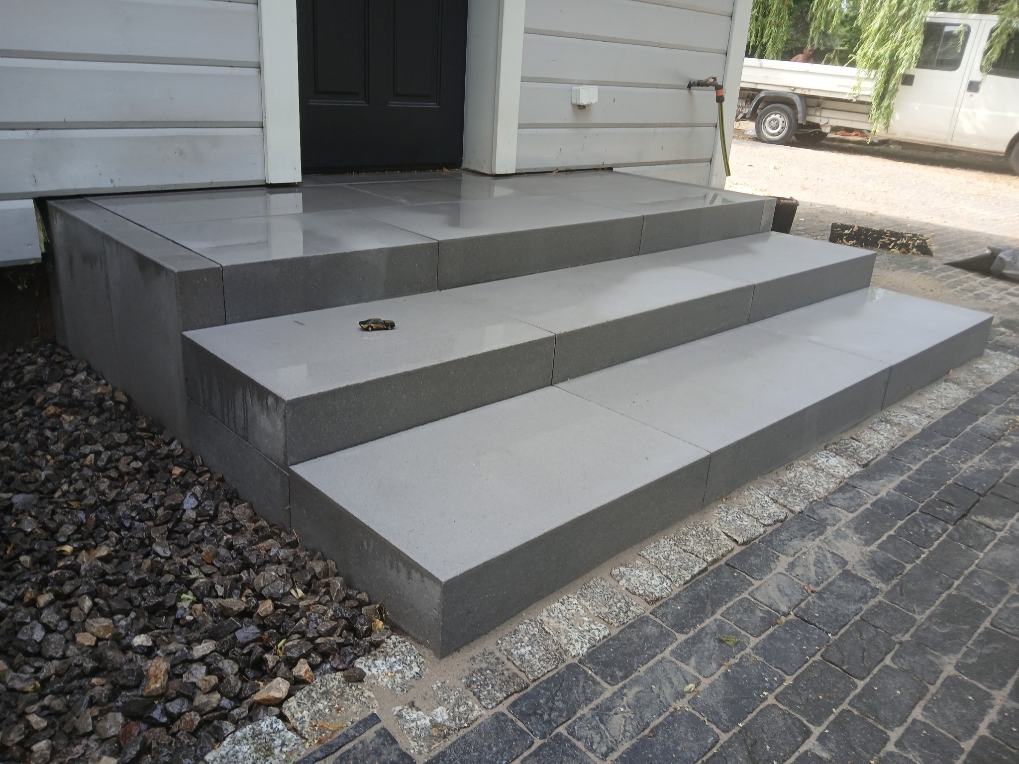 Trzy stopnie schodów wejściowych wyłożone szarą kostką brukową, z czarnymi drzwiami i ozdobną kostką na chodniku, zabawkowy samochód na drugim stopniu.