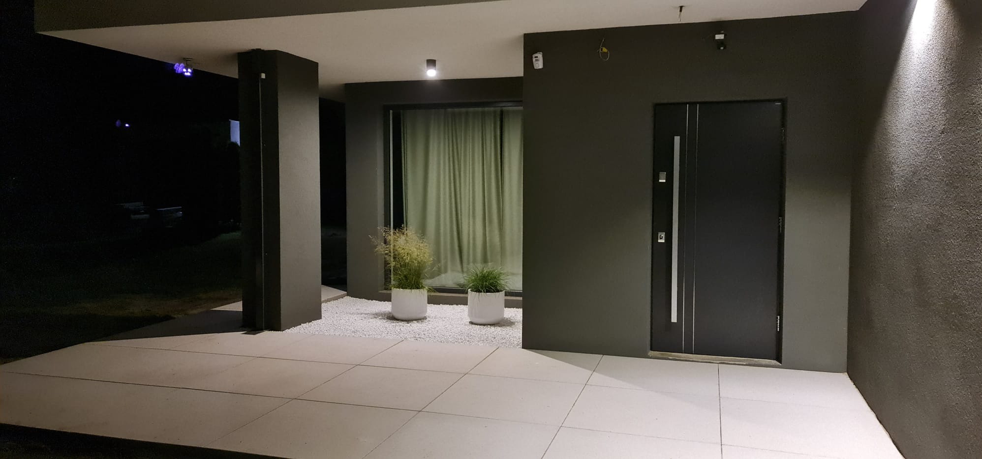 Nowoczesne wejście do domu z minimalistycznym ogrodem: dwie białe donice z trawami ozdobnymi na białym żwirze, szara elewacja i drzwi wejściowe, oświetlenie punktowe.