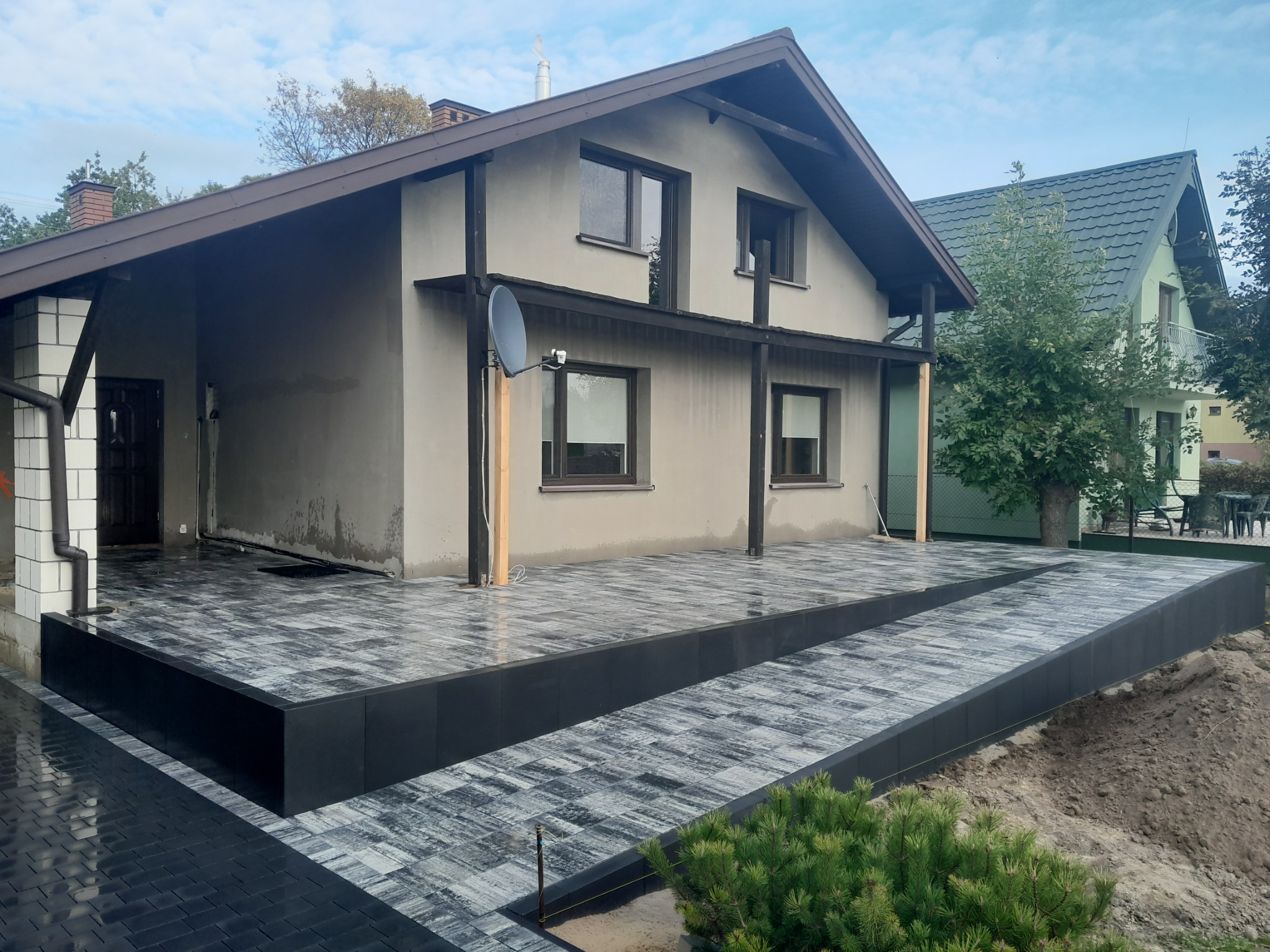Nowoczesny taras z szarej kostki brukowej ułożonej dwupoziomowo przed domem z brązowym dachem, widoczna antena satelitarna na elewacji budynku.