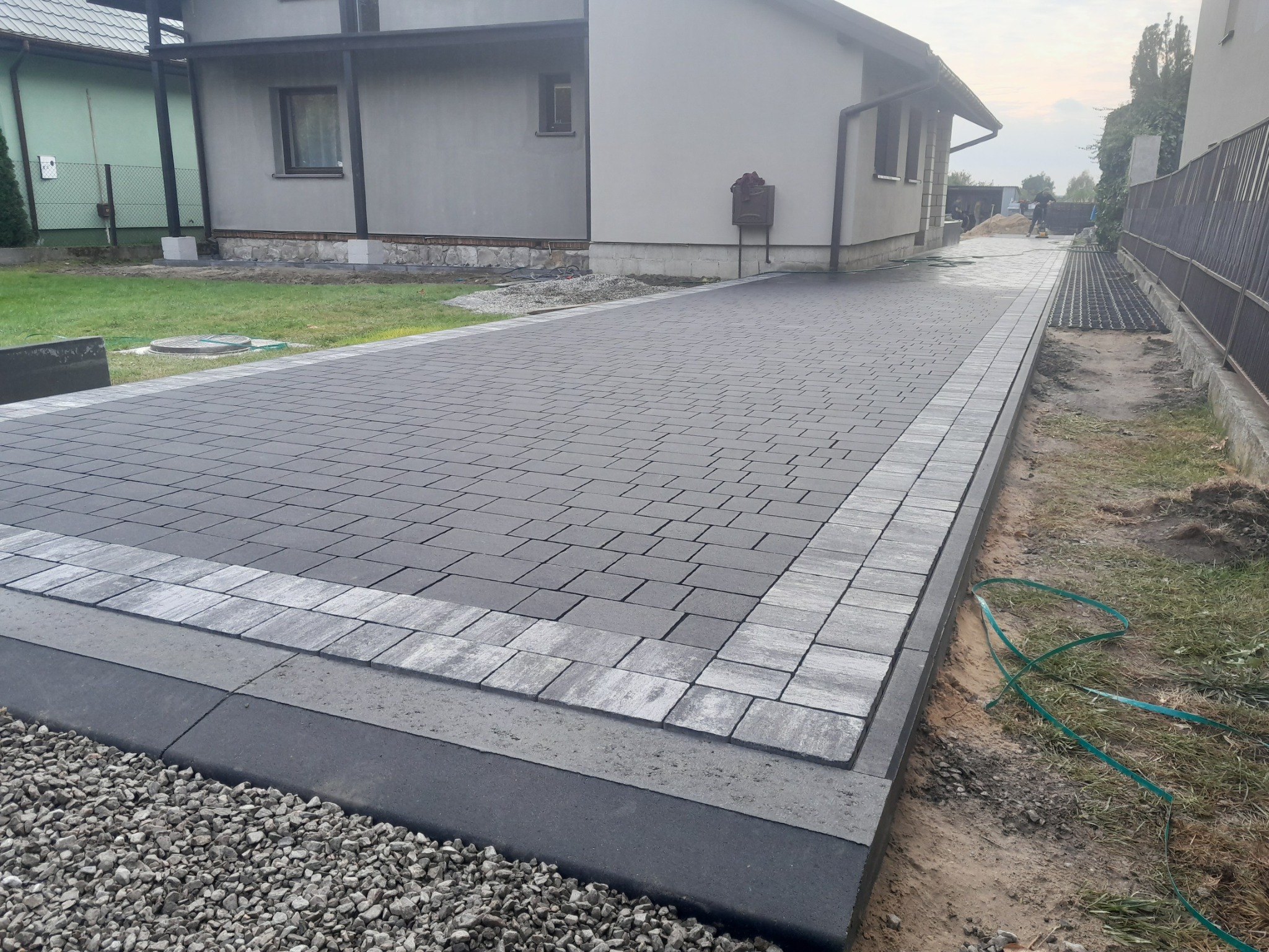 Nowo ułożony podjazd z szarej kostki brukowej w geometryczny wzór, obrzeża z jasnoszarej kostki, widoczne przygotowanie terenu pod trawnik, dom w tle.