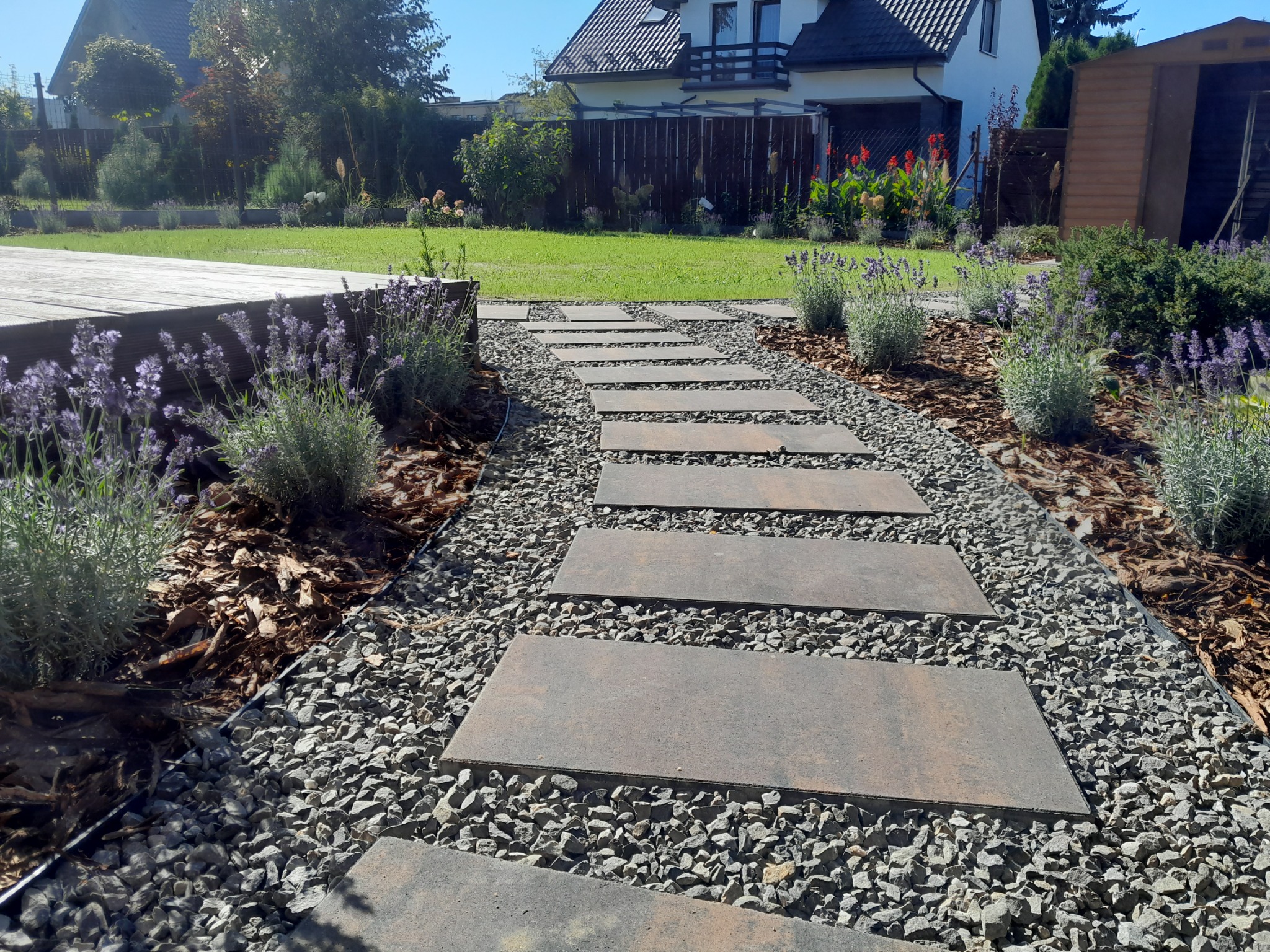 Ścieżka z prostokątnych płyt ułożona na żwirze, obrzeża z kory i lawendy, trawnik, dom jednorodzinny w tle.