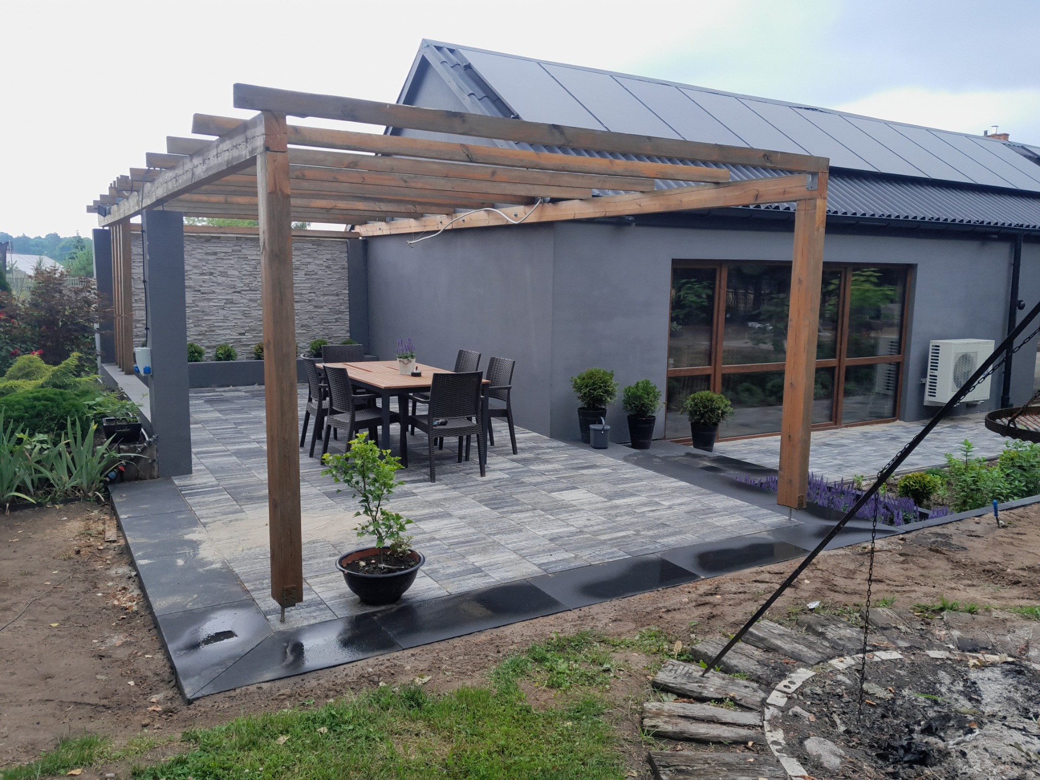 Nowoczesny taras z drewnianą pergolą, szarą kostką brukową i elementami zieleni, w tle szary budynek z oknami i kompresorem klimatyzacji.