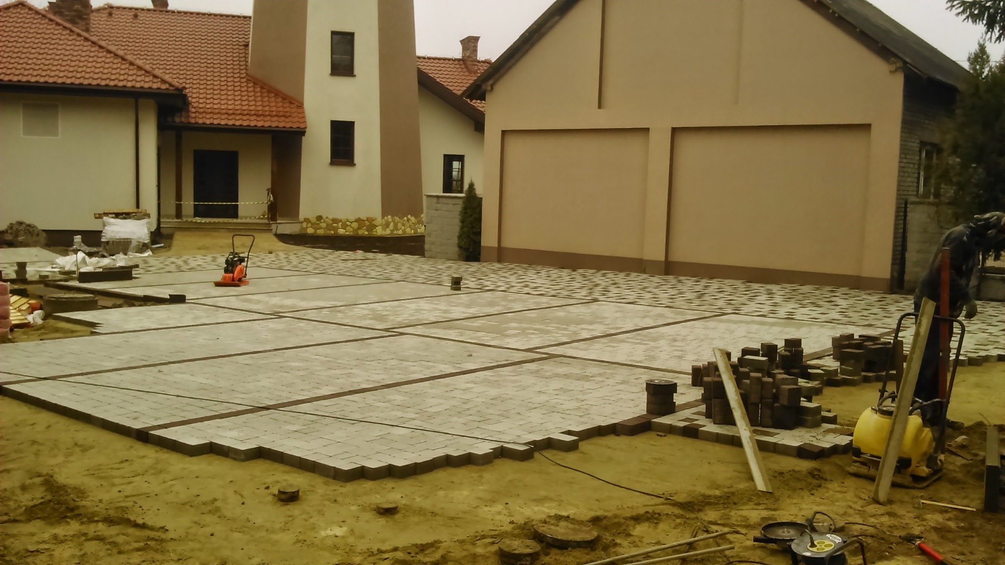 Fragment placu z częściowo ułożoną kostką brukową w odcieniach szarości i brązu, widoczny zagęszczarka wibracyjna, stos kostki oraz narzędzia, w tle budynki mieszkalne z czerwonymi dachami.