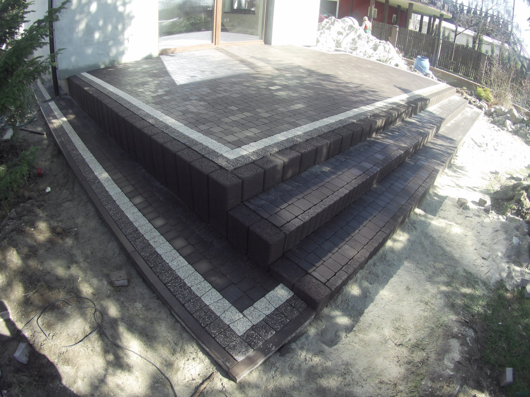 Schody i taras wyłożone brązową kostką brukową z szarym obramowaniem, widok z góry na świeżo ułożoną nawierzchnię, w tle dom z białymi ścianami i częściowo widocznym ogrodem.