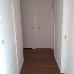 Korytarz z trzema białymi drzwiami z złotymi klamkami, brązowa podłoga z paneli, białe ściany, widok z perspektywy podłogi.