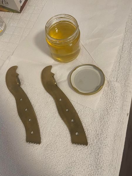 Słoik z bursztynowym płynem, otwarte wieczko i dwa metalowe narzędzia z rzędami otworów na białym ręczniku.